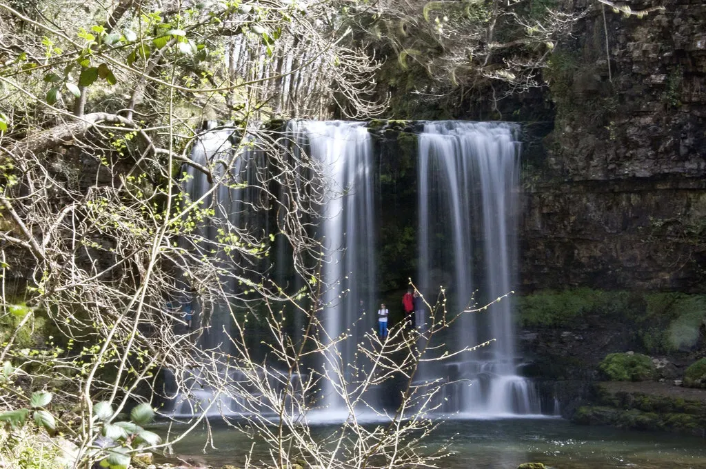 Sgwd yr Eira's wide curtain of falling water with walkers standing on the ledge behind it, framed by early spring branches